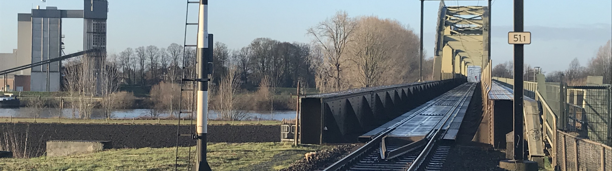 Stukje Maaslijn met spoorbrug rivierlandschap met industrieel gebouw en winterse bomen op de achtergrond