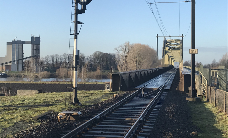 Stukje Maaslijn met spoorbrug rivierlandschap met industrieel gebouw en winterse bomen op de achtergrond