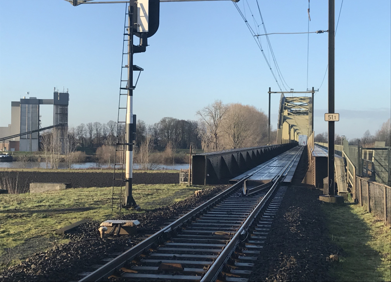 Stukje Maaslijn met spoorbrug rivierlandschap met industrieel gebouw en winterse bomen op de achtergrond