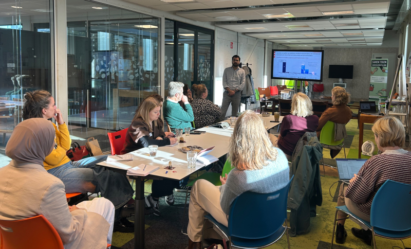 Deelnemers van de startbijeenkomst tijdens een interactieve werksessie, zittend rond een tafel luisteren naar een presentatie in een lichte, moderne vergaderruimte.
