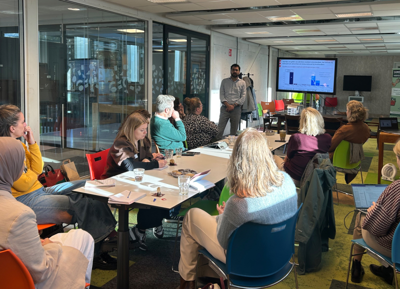 Deelnemers van de startbijeenkomst tijdens een interactieve werksessie, zittend rond een tafel luisteren naar een presentatie in een lichte, moderne vergaderruimte.