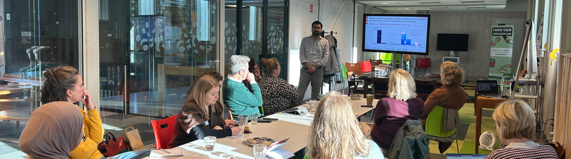 Deelnemers van de startbijeenkomst tijdens een interactieve werksessie, zittend rond een tafel luisteren naar een presentatie in een lichte, moderne vergaderruimte.
